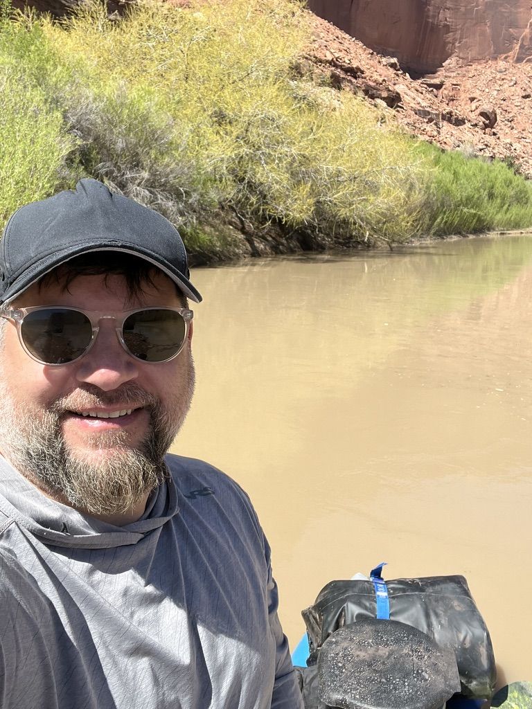 Karl, known as Quad, smiling in a cap and sunglasses on a muddy desert river below red rock