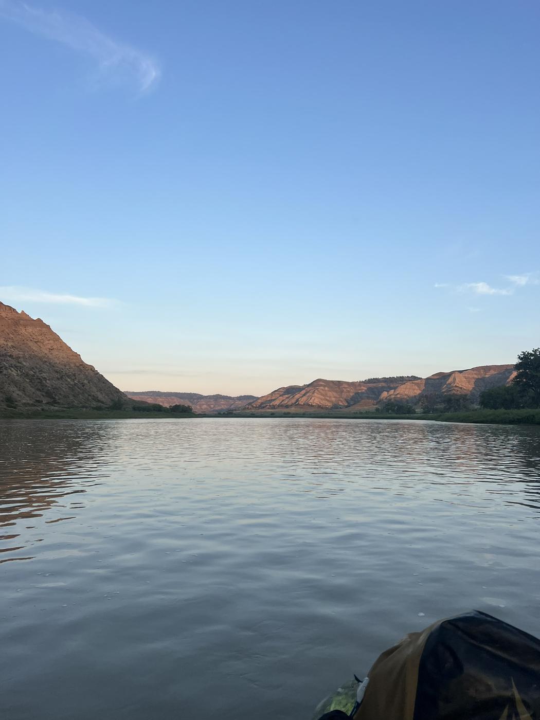 Early morning light on badlands hills across a wide, calm river, seen from the bow of a kayak