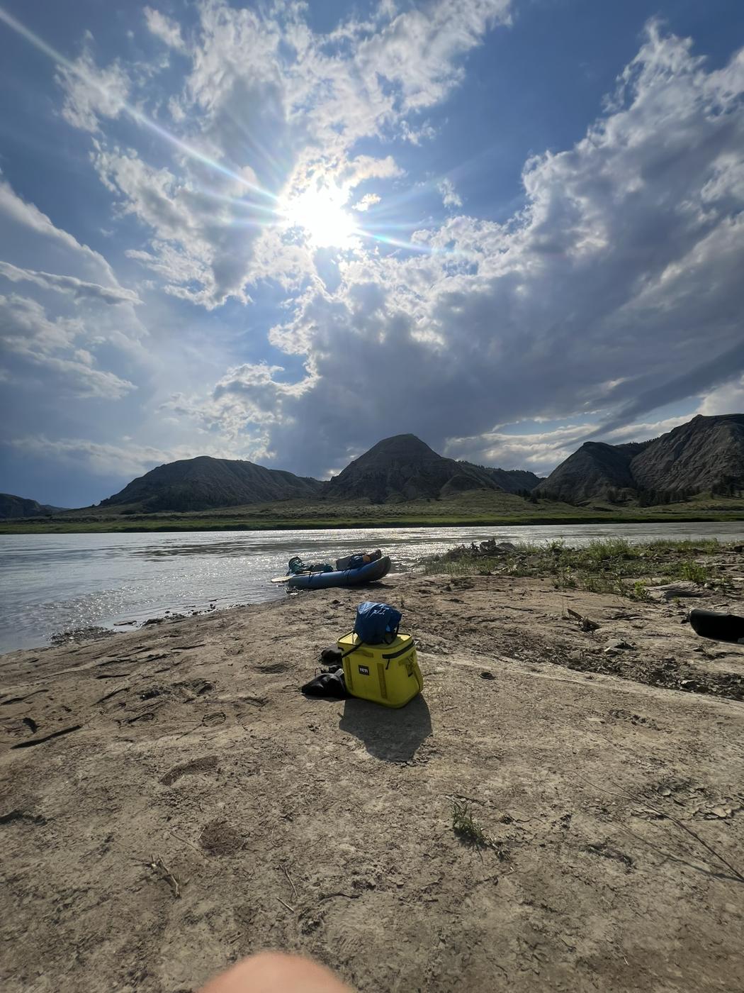 A cooler on a sandy beach with a loaded kayak at the water's edge and badlands peaks behind
