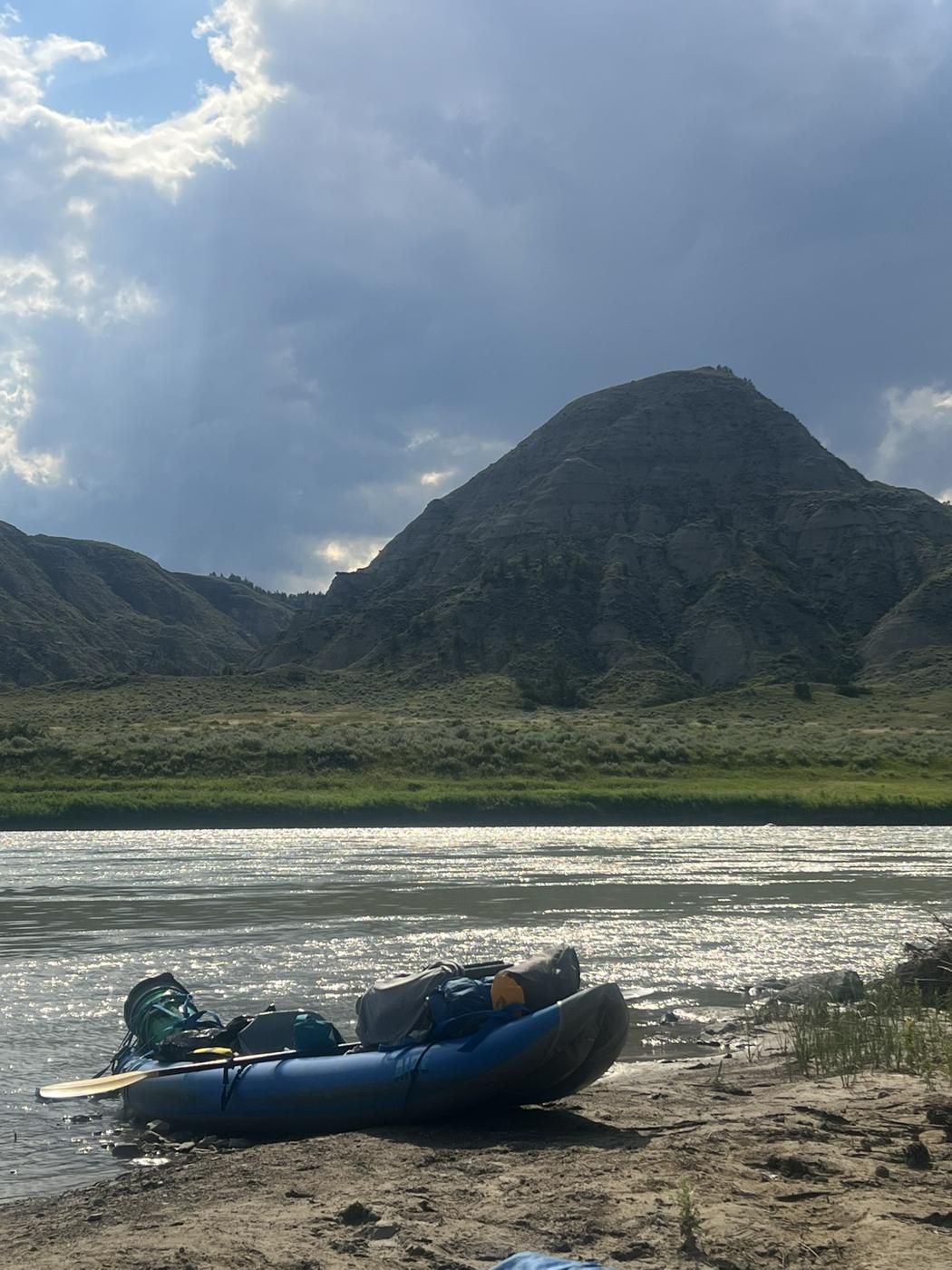 A loaded inflatable kayak on the bank beneath a dark badlands peak and storm light