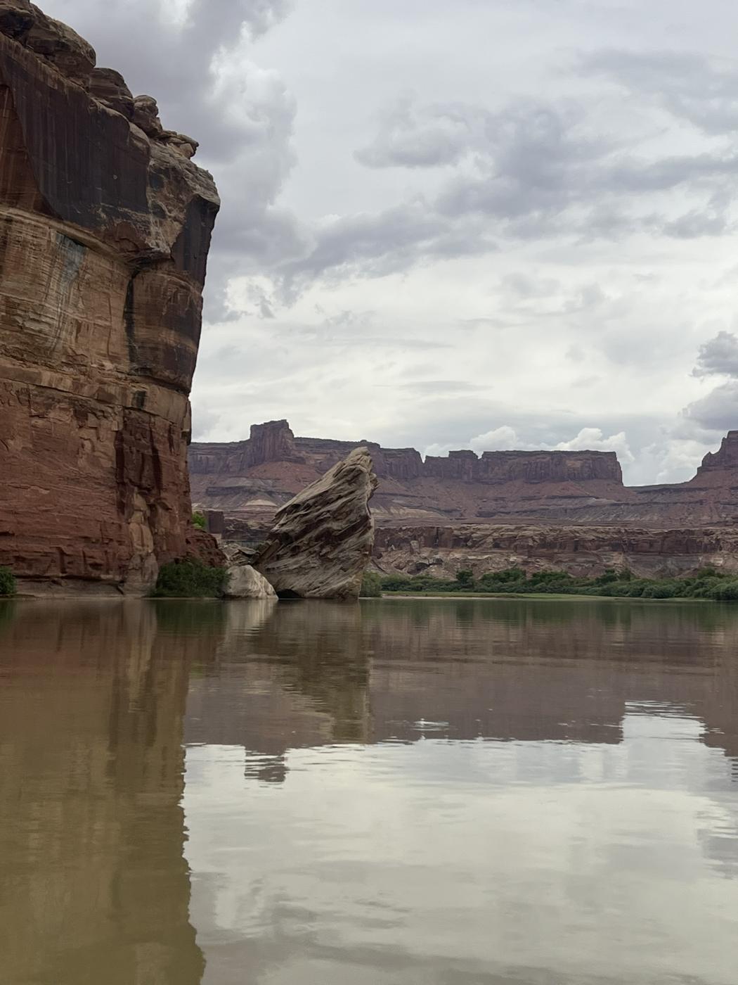 A tilted sandstone boulder in still brown water beneath red canyon walls and mesas